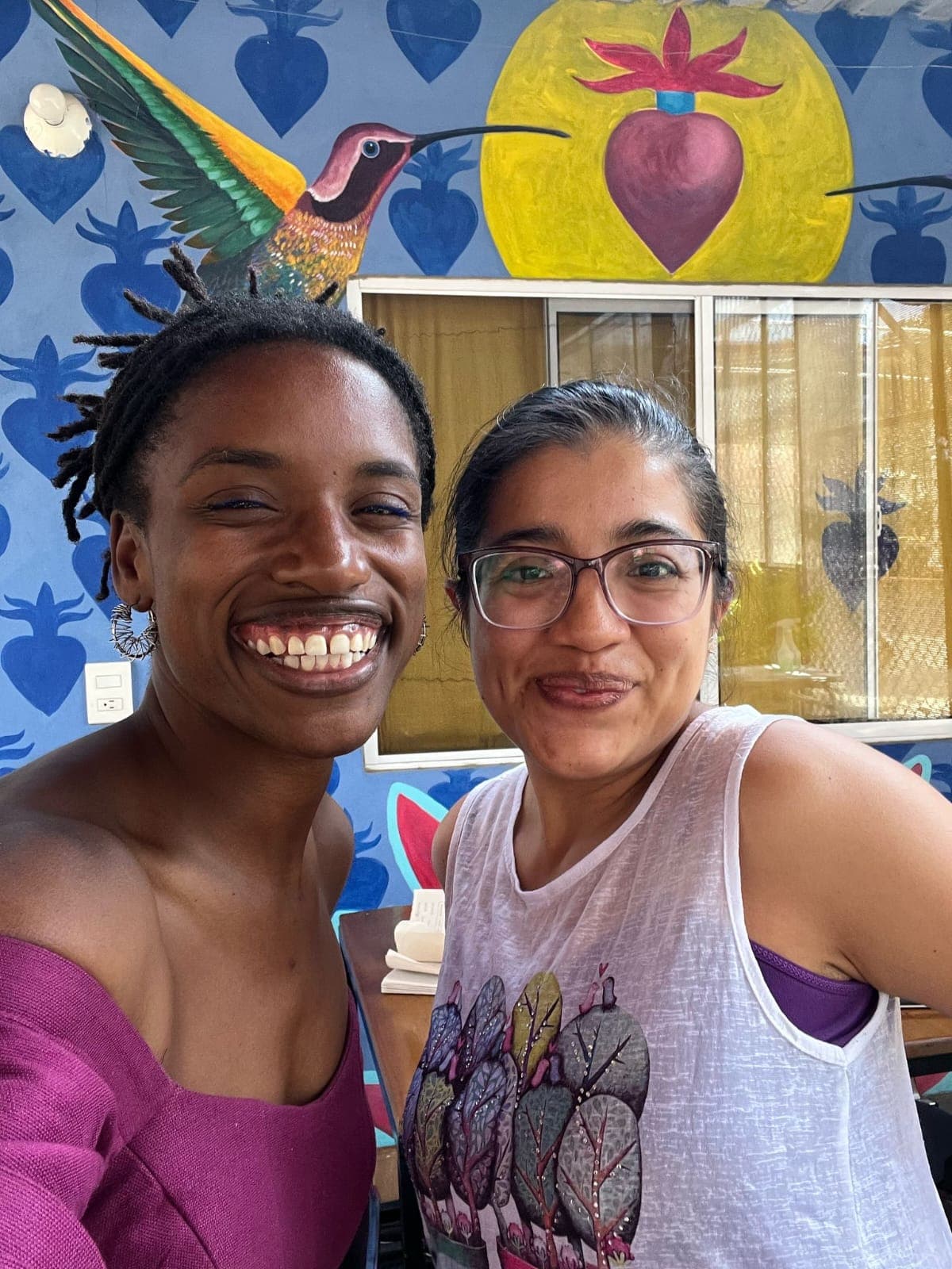 Spanish teacher and student smiling in front of a hand-painted hummingbird mural in the Oaxaca classroom